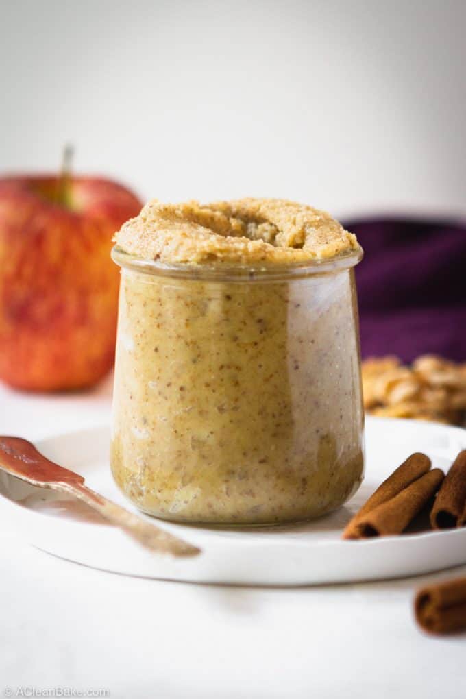 Small jar of apple cinnamon walnut butter sitting on a plate with a knife and cinnamon sticks and an apple in the background