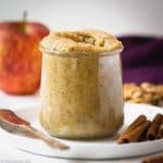 Small jar of apple cinnamon walnut butter sitting on a plate with a knife and cinnamon sticks