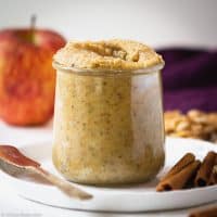 Small jar of apple cinnamon walnut butter sitting on a plate with a knife and cinnamon sticks