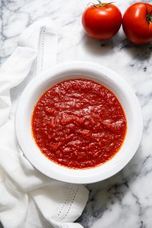 Bowl of simple pizza sauce resting on a white napkin and marble table