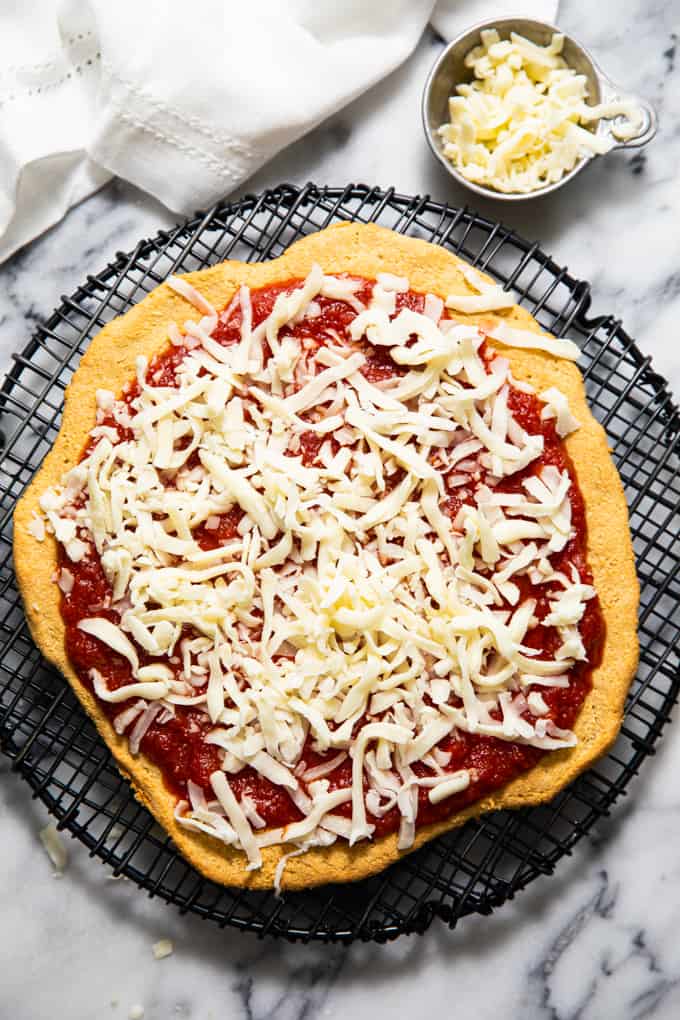 Gluten free paleo pizza crust topped with sauce and cheese on a rack before baking