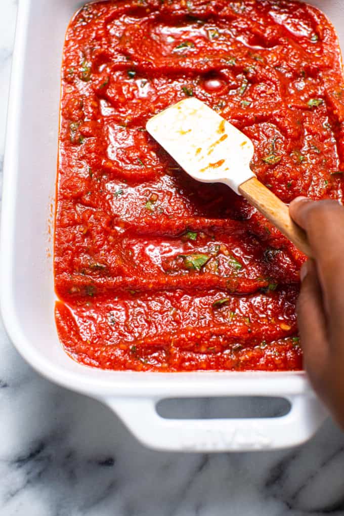 Tomato sauce being spread into a white casserole dish with white spatula