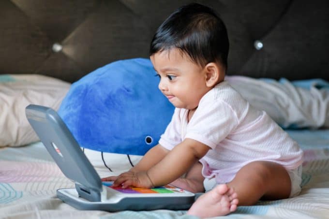 baby sitting up in a bed playing with a toy laptop - how to work from home with kids